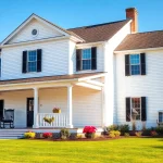 Explore colonial home construction with a charming white house featuring black shutters amidst a beautiful landscape.